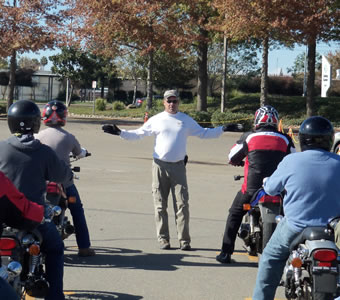 A group of motorcycle students lined up on their motorcycles listening to a safety lesson from the instruction at the head of the class
